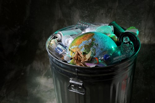 A metallic grey rubbish bin filled with various types of waste items, including crushed and flattened plastic bottles, transparent and green-tinted bottles, some with caps on and others without, along with crumpled brown paper and a small representation of the Earth globe made of plastic or similar material. The bin is situated outdoors on a concrete or paved surface, with a dark, slightly textured wall behind it, and is partially shaded, suggesting a typical setting for waste collection or disposal. The scene reflects general rubbish collection practices relevant to commercial or household waste removal in Lewisham or nearby areas, with the focus on recyclable plastic waste. The bin's metal surface has a handle and a locking mechanism at the front, indicating it is designed for secure and hygienic waste containment, and the plastic waste content demonstrates common household or office rubbish collection items. The lighting emphasizes the contents in the bin, highlighting the textures and colors of the bottles and paper, consistent with real waste disposal environments.