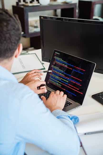 A person sitting at a desk in an office environment is working on a laptop with a dark screen displaying colorful lines of programming code. The individual, wearing a light blue shirt, is holding a pe