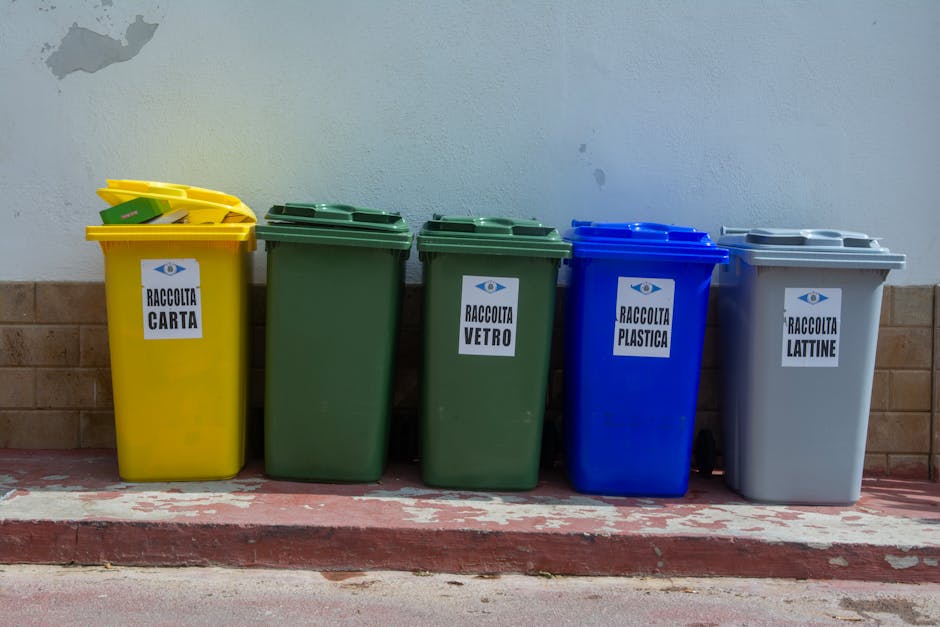 A row of five large, upright plastic waste bins positioned on a concrete curb against a pale blue wall with a brown brick base. From left to right, the bins are yellow, green, green, blue, and gray, e