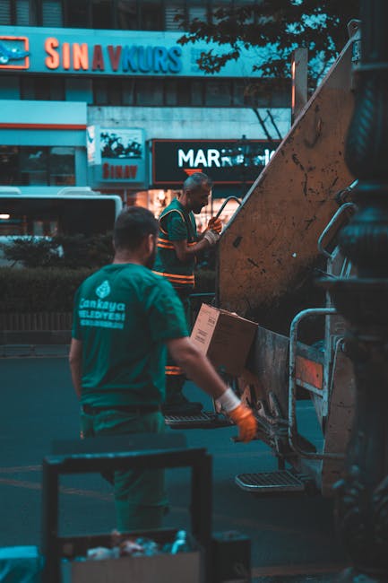 A collection of overflowing rubbish bins and scattered waste bags on a paved urban sidewalk, located in front of a building with a blue scaffolding structure and a shopfront. The large metal recycling