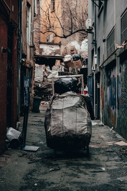 A large collection of mixed household and office rubbish is piled on a paved pavement area in front of a building, including a metal recycling bin overflowing with flattened cardboard boxes, white pla